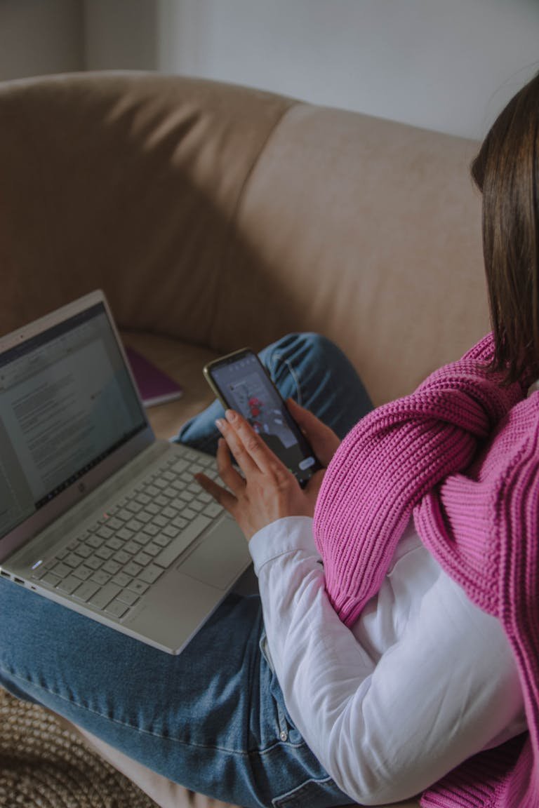 Woman sitting on a sofa multitasking with a smartphone and a laptop indoors.